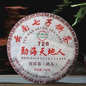 Peut inclure: Une galette de thé circulaire enveloppée dans du papier blanc avec des caractères chinois rouges et du texte anglais incluant Yun Nan Seven Tea Cake, 726, Meng Hai Heaven Earth, et un poids net de 357 grammes imprimé sur l'emballage.