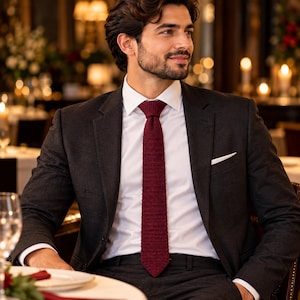 Puede incluir: Una chaqueta de traje gris carbón oscuro sobre una camisa blanca de cuello rígido, con una corbata de punto color burdeos intenso con un patrón tejido texturizado y un pañuelo blanco en el bolsillo izquierdo del pecho de la chaqueta.