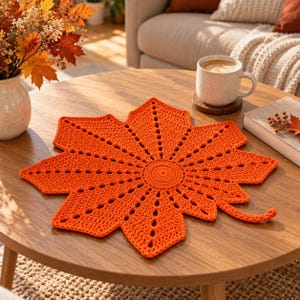 Pode incluir: Um centro de mesa de crochê laranja brilhante em forma de uma grande folha de bordo. A peça apresenta um centro circular com padrões rendados radiantes que formam as nervuras e os lóbulos pontiagudos, terminando num pequeno caule curvo.