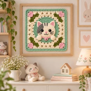 May include: A square crochet wall hanging featuring a central cat face with grey and white fur, pink ears, and a pink collar with a bell. The cat is framed by a green circle and a decorative border of cream, green, and pink floral crochet patterns.