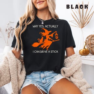 Può includere: Una t-shirt nera a maniche corte con una grafica arancione di una strega su una scopa con un gatto e una lanterna. Il testo recita: WHY YES, ACTUALLY I CAN DRIVE A STICK. La maglietta ha un girocollo ed è realizzata in tessuto nero.