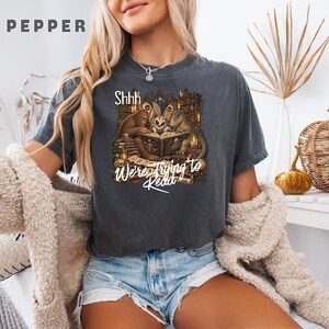 Può includere: Una t-shirt a maniche corte grigio antracite con una stampa grafica di Bigfoot, un gufo e un dinosauro che leggono un libro in una biblioteca. Il testo recita Shhh We're Trying to Read. La maglietta ha un girocollo e una vestibilità comoda.