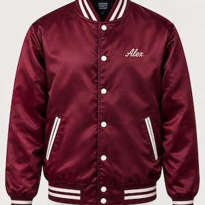 May include: A burgundy satin varsity jacket featuring white striped ribbed trim on the collar, cuffs, and hem. The jacket has a front snap-button closure and the name Alex embroidered in white script on the left chest. A label reads Custom Varsity Jacket.