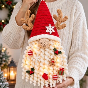 May include: A crocheted gnome ornament featuring a tall red hat with a white snowflake, tan reindeer antlers, and a long curly white beard. The beard is decorated with small crocheted ornaments including a gingerbread man, red stocking, gift, and holly.