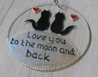 May include: A round glass ornament with a silver background and two black cats silhouetted on a hill. The cats are facing each other and there are three red hearts on the ornament. The text "Love you to the moon and back" is written in black paint on the ornament. The text "Copyright Joanne Simpson" is written in small black letters at the bottom of the ornament.