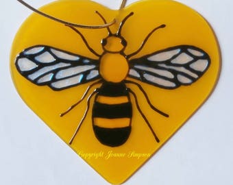May include: A yellow heart-shaped glass ornament with a black and yellow bee painted on it. The bee has white wings with black veins. The ornament has a gold string for hanging. The text "Copyright Joanne Simpson" is printed on the bottom of the ornament.