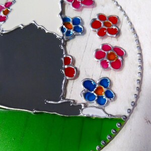 May include: A round, stained glass ornament with a gray and white silhouette of a sheep surrounded by colorful flowers. The flowers are red, pink, blue, and orange. The ornament has a green background and a silver border with small dots. The text "Copyright Joanne Simpson" is at the bottom of the ornament.