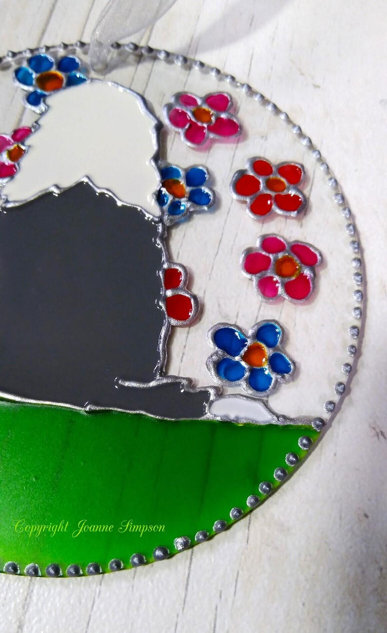 May include: A round, stained glass ornament with a gray and white silhouette of a sheep surrounded by colorful flowers. The flowers are red, pink, blue, and orange. The ornament has a green background and a silver border with small dots. The text "Copyright Joanne Simpson" is at the bottom of the ornament.