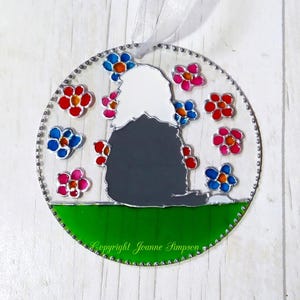 May include: A round stained glass ornament featuring a black and white dog silhouette surrounded by colorful flowers. The dog is sitting on a green field with the text "Copyright Joanne Simpson" at the bottom.