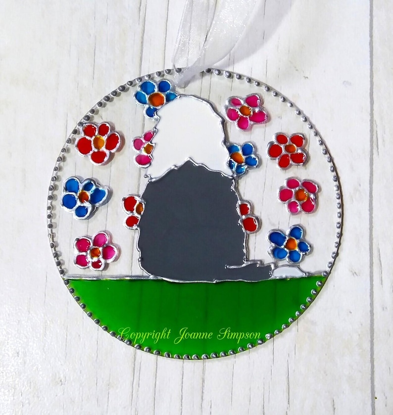 May include: A round stained glass ornament featuring a black and white dog silhouette surrounded by colorful flowers. The dog is sitting on a green field with the text "Copyright Joanne Simpson" at the bottom.