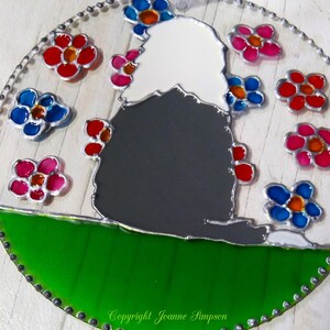 May include: A stained glass ornament featuring a black and white dog sitting on a green hill with a white background. The dog is surrounded by colorful flowers in shades of pink, red, blue, and orange. The ornament is framed with a silver border and has a silver chain for hanging. The text "Copyright Joanne Simpson" is visible at the bottom of the ornament.