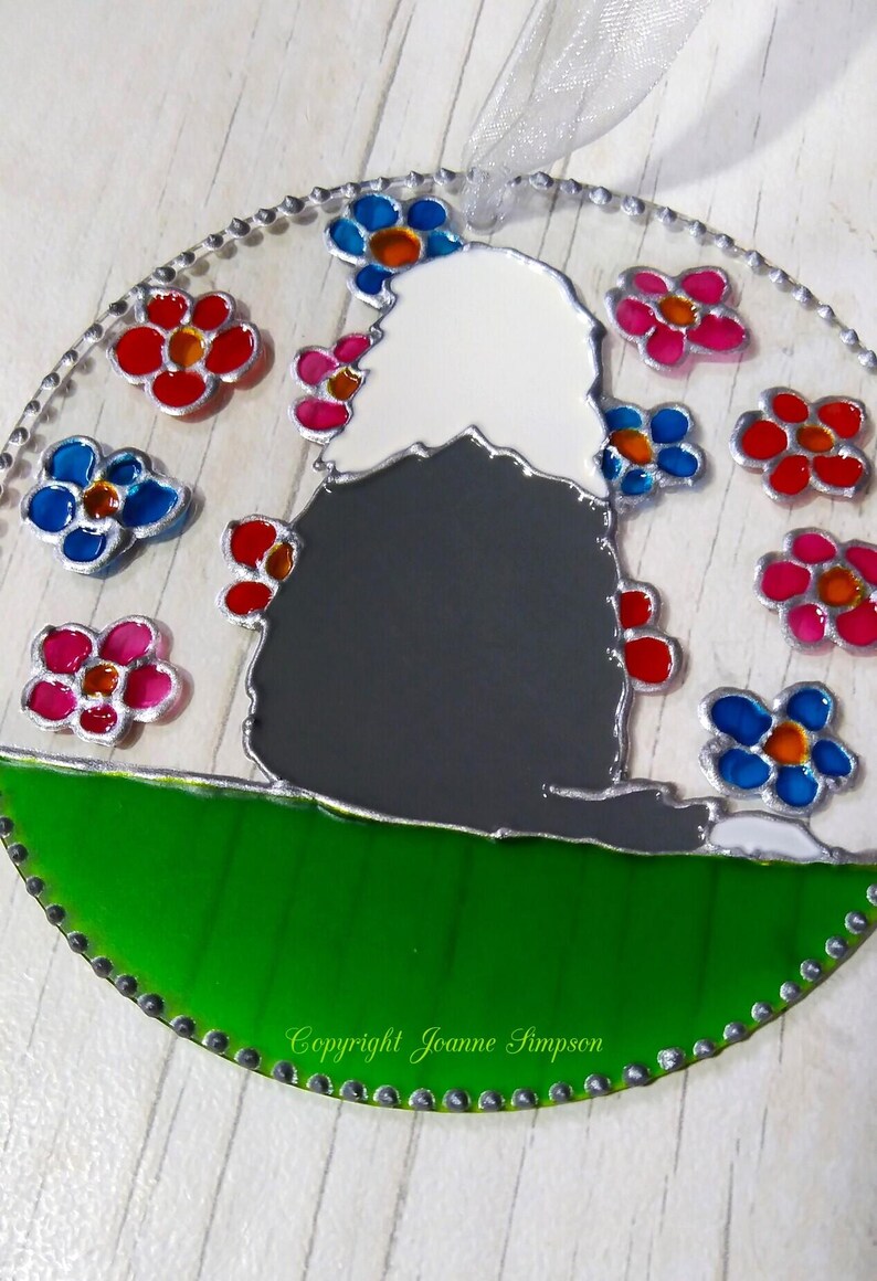 May include: A stained glass ornament featuring a black and white dog sitting on a green hill with a white background. The dog is surrounded by colorful flowers in shades of pink, red, blue, and orange. The ornament is framed with a silver border and has a silver chain for hanging. The text "Copyright Joanne Simpson" is visible at the bottom of the ornament.