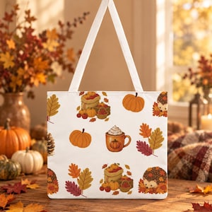 May include: A white tote bag featuring a repeating autumn-themed pattern. The design includes orange pumpkins, brown hedgehogs nestled in fall leaves, mugs of hot cocoa with whipped cream, and burlap sacks filled with various seasonal produce.