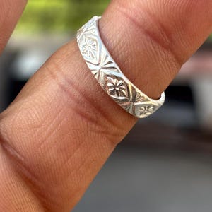 Peut inclure: Une bague en argent ornée d'un motif gravé répétitif de petites fleurs à six pétales inscrites dans des losanges. L'anneau présente une finition métallique polie et une largeur constante, mettant en valeur des détails floraux symétriques.