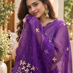 May include: A vibrant purple sheer saree featuring delicate gold embroidery and scalloped edges. The fabric is adorned with scattered gold floral motifs and a larger, intricate geometric floral pattern on the lower section, paired with gold jhumka earrings.