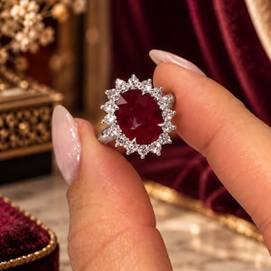 Puede incluir: Un anillo de tono plateado con un gran rubí rojo oscuro de corte ovalado en el centro, rodeado por un halo de pequeñas piedras blancas redondas y transparentes. El engaste es metálico y pulido, con un diseño clásico de racimo.