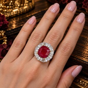 Puede incluir: Un anillo que presenta una gran piedra preciosa de color rojo intenso con talla cojín en el centro, rodeada por un halo de numerosas piedras blancas pequeñas, claras y con talla brillante dispuestas en círculo sobre una banda metálica.