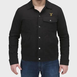 Puede incluir: Una chaqueta negra con botones, cuello en punta, mangas largas y un bolsillo en el pecho con solapa. Un logotipo amarillo con la letra Y está bordado en la parte superior izquierda del pecho. La chaqueta se lleva sobre vaqueros azules.