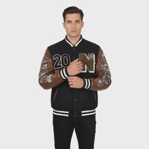 Op de afbeelding: Een zwart varsity-jack met bruine mouwen, voorzien van wit gestreepte geribbelde boorden en kraag. De voorzijde toont het getal 20 en de letter N met honkbalstiksels. Beide mouwen zijn versierd met diverse geborduurde patches.