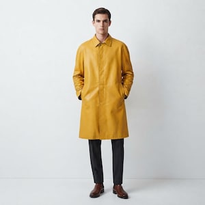 Puede incluir: Un abrigo color amarillo mostaza hasta la rodilla con cuello en punta, mangas largas y una tapeta de botones oculta en la parte delantera. El abrigo se lleva sobre pantalones gris carbón oscuro y zapatos de vestir de cuero marrón.