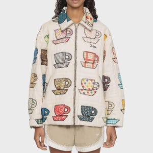 Puede incluir: Una chaqueta acolchada de color crema con cremallera que presenta un patrón repetitivo de varias tazas de té en diferentes colores y estampados de tela. La chaqueta tiene cuello en punta, mangas largas y la palabra Sea bordada en negro.