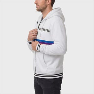 May include: A white zip-up hoodie featuring a hood with drawstrings, long sleeves with black striped cuffs, and a black front zipper. The chest area displays a horizontal graphic pattern consisting of thin black lines above a blue, purple, and red stripe.
