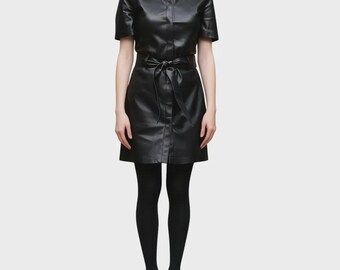 Peut inclure: Une robe chemise en similicuir noir à manches courtes, dotée d'un col classique et d'une fermeture boutonnée sur le devant. La robe comprend une ceinture à nouer assortie à la taille, créant une silhouette cintrée. Portée avec des collants noirs opaques.