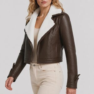 May include: A dark brown faux leather jacket featuring a wide white sherpa collar and lapels. The jacket has long sleeves with decorative cuffs, a cropped length, and a smooth finish. It is worn over a light beige top and light-colored pants.