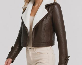 Peut inclure: Une veste en similicuir marron foncé dotée d'un large col et de revers en sherpa blanc. La veste présente des manches longues avec des poignets décoratifs, une coupe courte et une finition lisse. Elle est portée sur un haut beige clair.