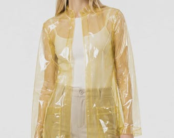 May include: A transparent yellow raincoat featuring a high collar, long sleeves, and two large square patch pockets on the front. The jacket is worn over a plain white top and light-colored trousers, showcasing a clear, glossy, and smooth finish.