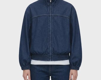 Peut inclure: Une veste en jean bleu foncé dotée d'une fermeture éclair intégrale sur le devant, d'un col montant et de manches longues avec poignets élastiqués. La veste présente une couture horizontale sur la poitrine et un ourlet élastiqué sur un t-shirt blanc.