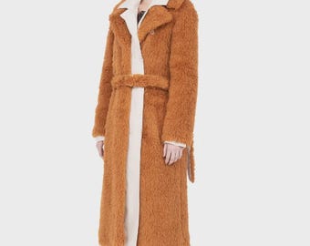 Peut inclure: Un long manteau texturé de couleur rouille avec une finition duveteuse semblable à de la fausse fourrure, un large col cranté et une ceinture en tissu assortie à la taille. Le manteau est porté sur un vêtement clair à la texture lisse.