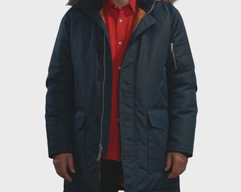 Peut inclure: Une parka bleu foncé avec une capuche bordée de fausse fourrure marron, une fermeture éclair frontale et deux grandes poches à rabat. La veste est portée sur une chemise rouge vif à boutons. Le modèle inclut une poche utilitaire sur la manche gauche.