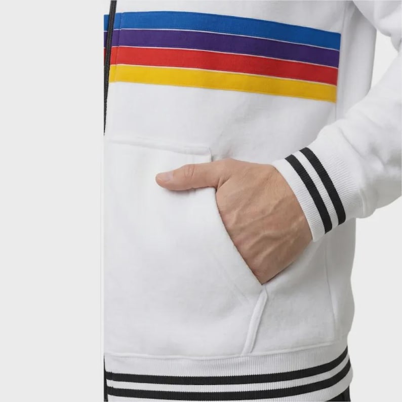 May include: A white zip-up jacket featuring horizontal stripes in blue, purple, red, and yellow across the chest. The garment includes a black zipper, a front pocket, and ribbed cuffs and hem with two black stripes on a white background.