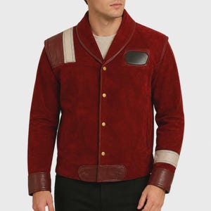 Può includere: Una giacca in camoscio rosso scuro con collo a scialle, bottoni a pressione color oro e dettagli in pelle liscia. Il design presenta una striscia verticale beige chiaro sulla spalla destra, una toppa rettangolare nera sul petto e polsini marrone scuro.
