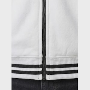 May include: A white zip-up jacket featuring a central black zipper closure. The garment includes a ribbed hem with two distinct black horizontal stripes running along the bottom edge, providing a clear contrast against the solid white fabric.