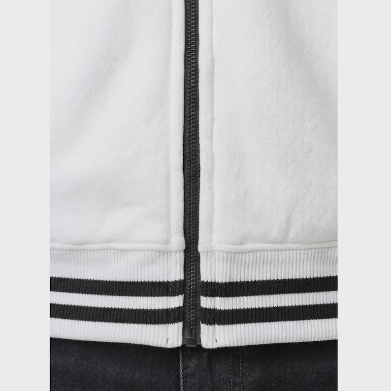 May include: A white zip-up jacket featuring a central black zipper closure. The garment includes a ribbed hem with two distinct black horizontal stripes running along the bottom edge, providing a clear contrast against the solid white fabric.