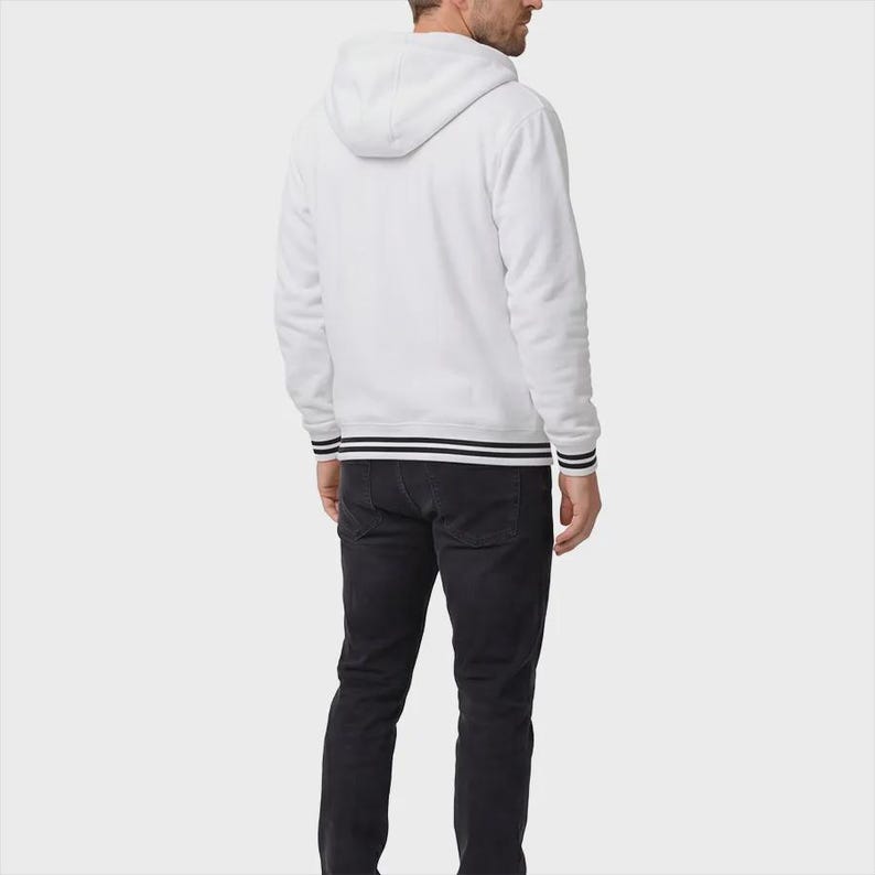 May include: A white hooded sweatshirt featuring long sleeves and a hood, paired with dark charcoal gray jeans. The sweatshirt has black and white striped ribbed trim along the cuffs and the bottom hem, providing a distinct contrast to the solid white fabric.