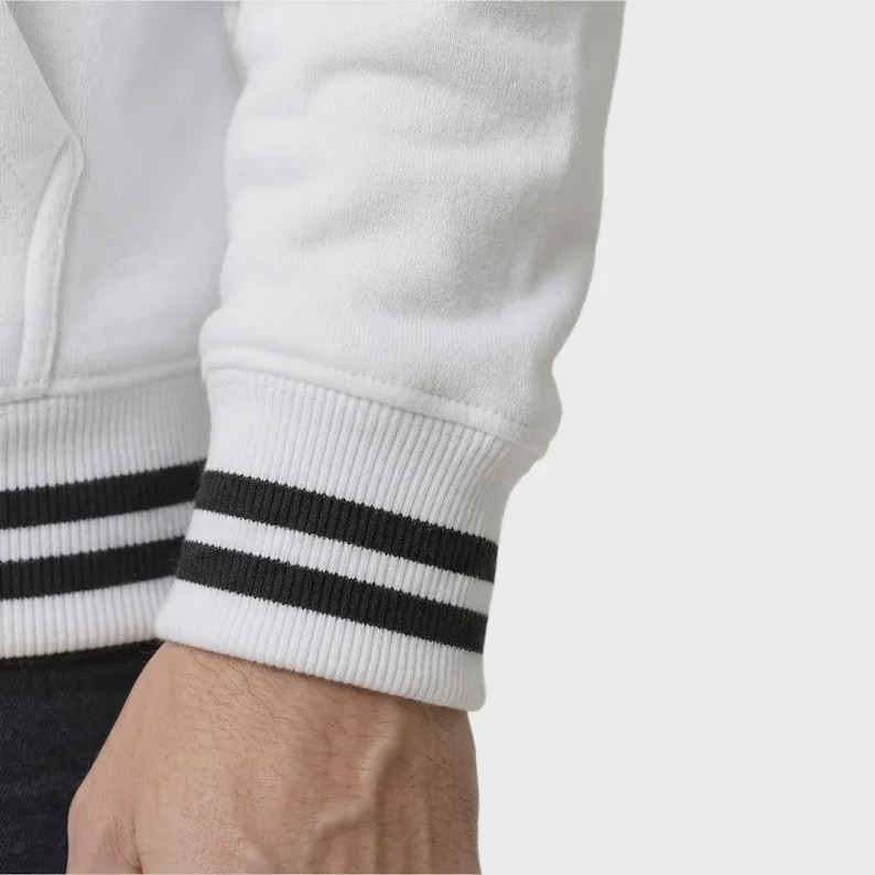 May include: A white long-sleeved sweatshirt featuring a ribbed cuff at the wrist. The cuff is white with two distinct horizontal black stripes running around the circumference, providing a contrasting detail against the solid white fabric of the sleeve.