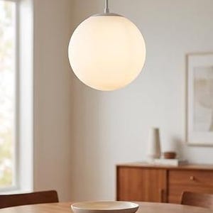 May include: A spherical pendant light fixture featuring a smooth, opaque white glass globe suspended from a thin, light-colored cord. The lamp emits a soft, diffused glow, hanging centrally above a round wooden dining table in a room.