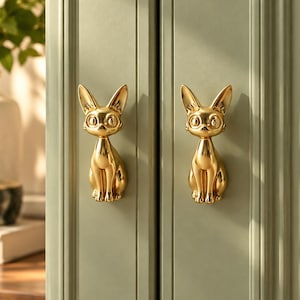 May include: Two gold-toned cabinet pulls shaped like sitting cats with pointed ears and large eyes, mounted on a sage green paneled cabinet door. The metallic cat figures feature a smooth, reflective finish and are positioned vertically.