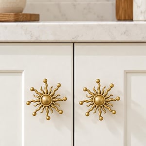 May include: Two brass-toned cabinet knobs shaped like suns with radiating wavy rays and small spherical tips. Each knob features a central circular disc surrounded by alternating long and short curved metal arms, mounted on white cabinet doors.