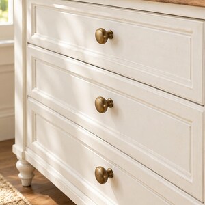 May include: A white wooden dresser featuring three horizontal drawers with recessed panel detailing. Each drawer is equipped with a single round, brushed brass knob. The dresser has a natural light brown wood top surface with a visible grain texture.
