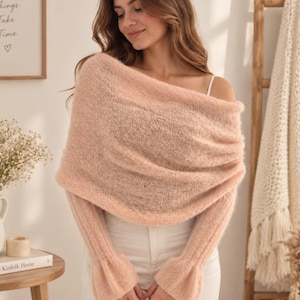 May include: A soft, pale peach-colored knitted off-the-shoulder wrap with long sleeves featuring flared cuffs. The garment is crafted from a lightweight, semi-sheer, fuzzy yarn with a loose, open-knit texture that drapes across the upper torso.