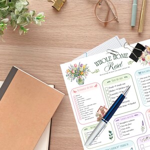 May include: A printed Whole Home Reset checklist featuring floral illustrations, checkboxes, and task lists for various rooms. The document is accompanied by a blue and silver ballpoint pen, a brown notebook, eyeglasses, and gold binder clips.