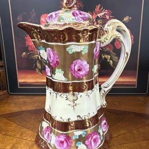 May include: A porcelain coffee pot featuring a metallic gold and deep maroon finish with pink rose floral patterns. The vessel includes a matching lid with a decorative finial, a curved handle, and a spout, all accented with gold trim details.