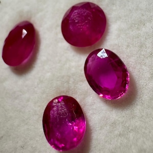 Puede incluir: Cuatro piedras preciosas facetadas de forma ovalada en un color magenta rosado intenso y vibrante. Cada piedra presenta una superficie pulida con múltiples facetas geométricas que reflejan la luz, creando brillos intensos en sus superficies.