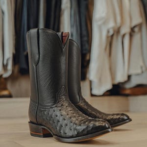 Puede incluir: Un par de botas de estilo western negras con un empeine de cuero texturizado con patrón de plumas y una caña de cuero granulado liso. Las botas tienen punta afilada, tacón apilado con una fina raya roja y una abertura en forma de V.