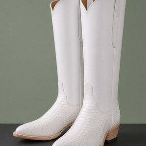 Puede incluir: Un par de botas altas blancas con una caña de cuero liso y una zona del pie con textura de escamas. Las botas tienen punta afilada, un tacón de madera apilado y una abertura profunda en forma de V en la parte superior de la caña.