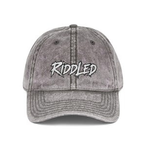 May include: A charcoal grey acid-wash baseball cap featuring the word RIDDLED embroidered in white with a jagged, dripping font style across the front panels. The cap has a curved brim, a button on top, and stitched eyelets for ventilation.